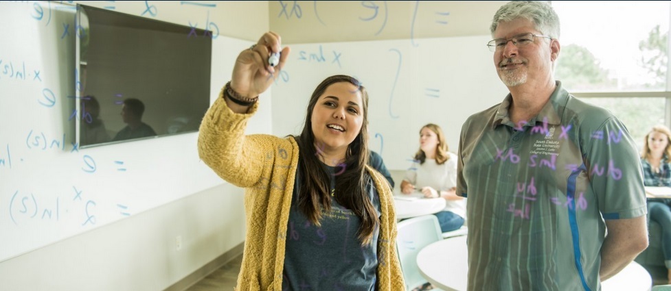 Student writing equations on a board with the professor standing next to her.